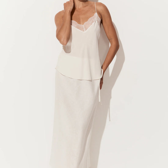 A woman modeling a two-piece white loungewear set consisting of a camisole top with lace trim and a matching long skirt, set against a plain white studio background.