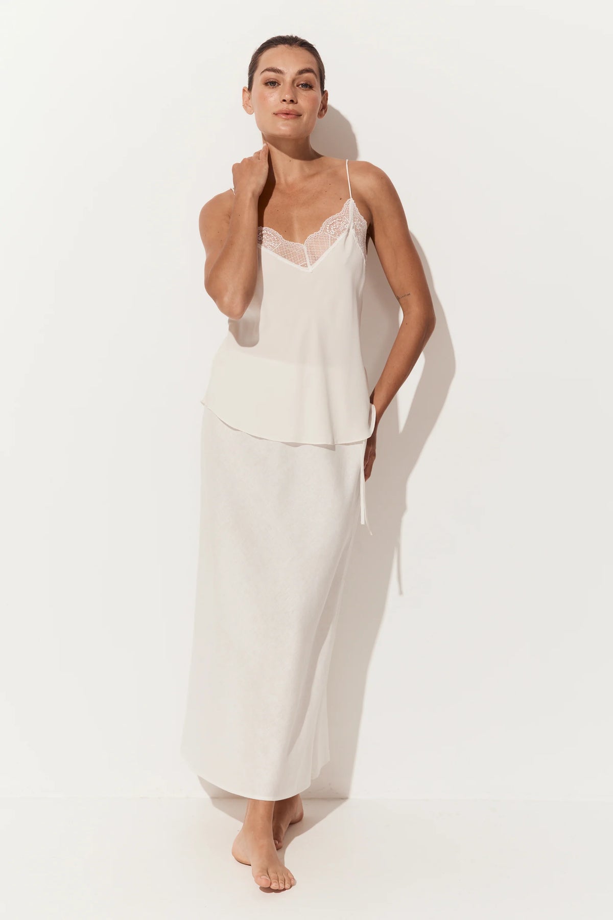 A woman modeling a two-piece white loungewear set consisting of a camisole top with lace trim and a matching long skirt, set against a plain white studio background.