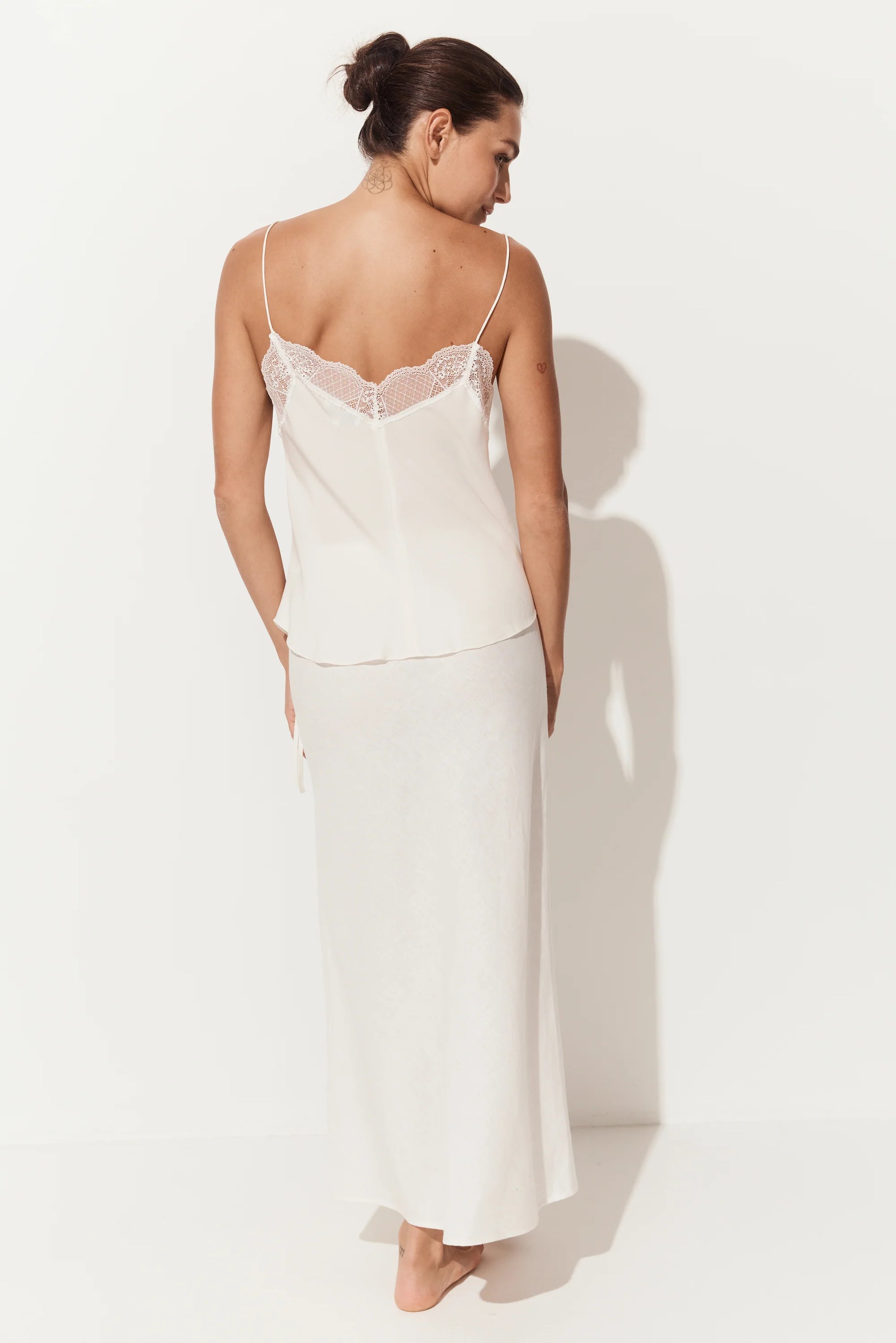 A woman shown from the back wearing a two-piece slip set consisting of a cream camisole with lace trim and a matching long skirt, set against a plain white studio background with a soft shadow.