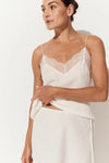 A woman wearing a white camisole top with thin straps, a scalloped lace neckline, and a draped wrap-style skirt, photographed against a plain light background.
