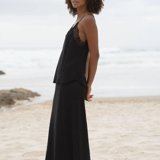 A woman with curly brown hair stands barefoot on a sandy beach wearing a black two-piece linen set consisting of a camisole top with sheer lace shoulder panels and a matching maxi skirt.