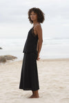 A woman with curly brown hair stands barefoot on a sandy beach wearing a black two-piece linen set consisting of a camisole top with sheer lace shoulder panels and a matching maxi skirt.