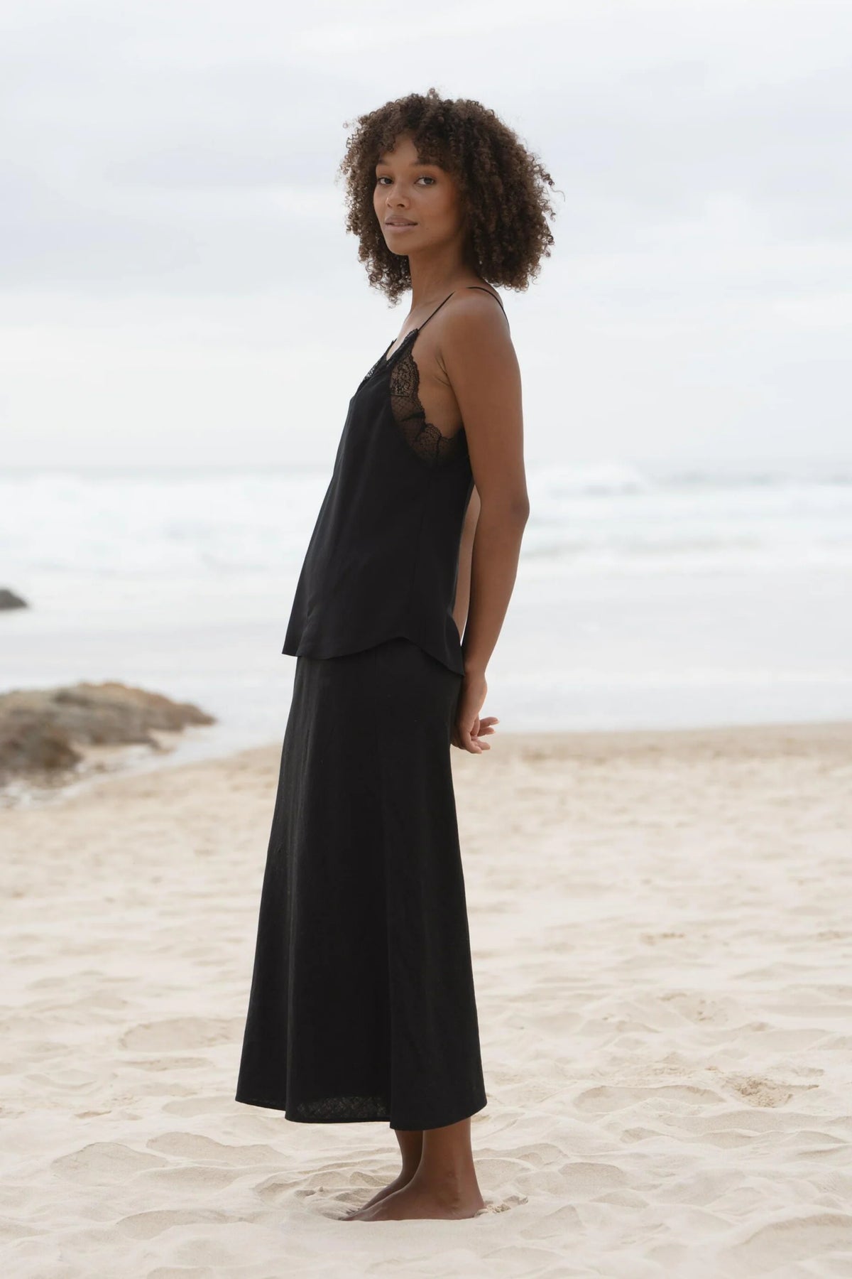 A woman with curly brown hair stands barefoot on a sandy beach wearing a black two-piece linen set consisting of a camisole top with sheer lace shoulder panels and a matching maxi skirt.