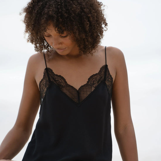 A woman with curly brown hair wearing a black camisole top with black lace trim at the neckline, photographed outdoors against a soft, bright sky background.
