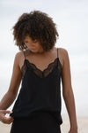 A woman with curly brown hair wearing a black camisole top with black lace trim at the neckline, photographed outdoors against a soft, bright sky background.