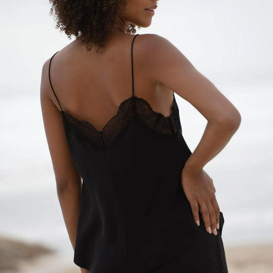 A woman with curly brown hair is shown from behind wearing a black camisole slip dress with a lace-trimmed open back, set against a soft-focus beach background.