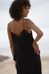A woman with curly brown hair is shown from behind wearing a black camisole slip dress with a lace-trimmed open back, set against a soft-focus beach background.