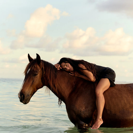 A woman in a black outfit rests her head on the neck of a brown horse standing in shallow water under a cloudy sky.