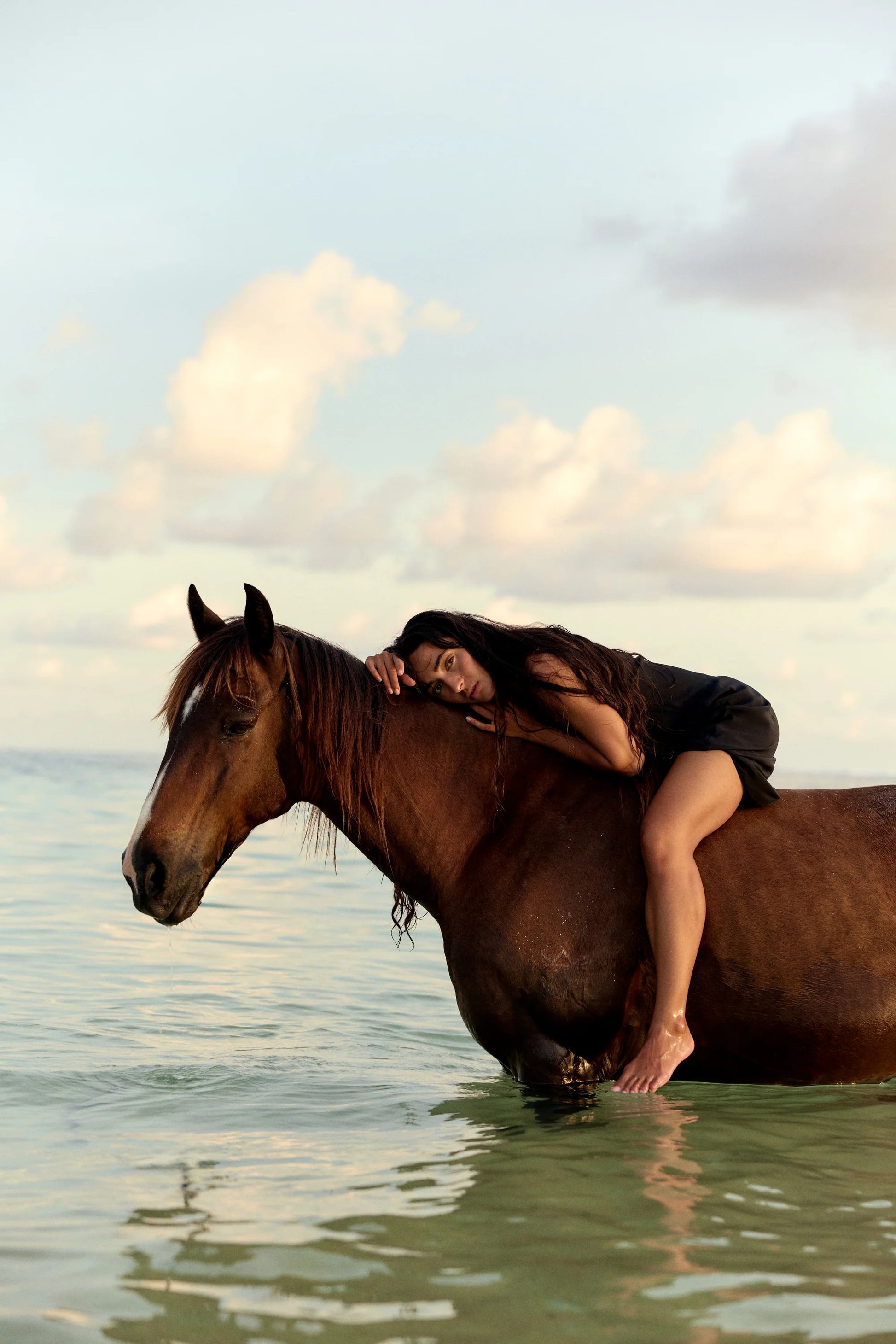 A woman in a black outfit rests her head on the neck of a brown horse standing in shallow water under a cloudy sky.