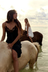 Two women posing with horses in shallow water at sunset. The woman in the foreground wears a black lace-trim camisole dress and sits on a light-colored horse, while the woman in the background wears a white dress and sits on a darker horse.