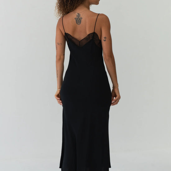 A woman stands barefoot against a plain light gray studio background, photographed from the back. She is wearing a black spaghetti-strap maxi dress with a sheer black lace upper back and shoulder detail.