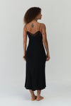 A woman stands barefoot against a plain light gray studio background, photographed from the back. She is wearing a black spaghetti-strap maxi dress with a sheer black lace upper back and shoulder detail.