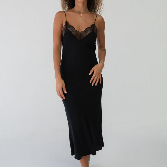 A woman modeling a black midi slip dress with thin straps and a lace V-neckline, standing against a plain white studio background.