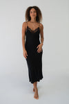 A woman modeling a black midi slip dress with thin straps and a lace V-neckline, standing against a plain white studio background.