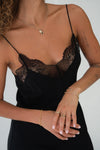 A close-up of a model wearing a black lace-trimmed camisole top with thin straps, paired with delicate gold jewelry against a plain light background.