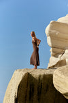 A woman in a sleeveless brown maxi dress stands barefoot on a large rock formation under a clear blue sky.