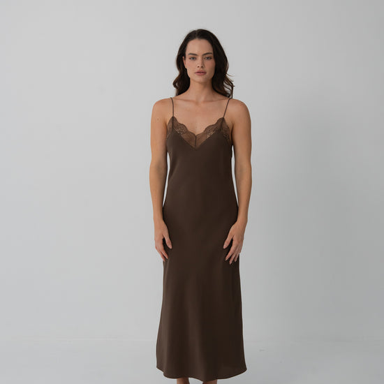 A woman modeling a dark brown slip dress with a lace V-neckline, standing against a plain light gray studio background.