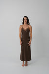 A woman modeling a dark brown slip dress with a lace V-neckline, standing against a plain light gray studio background.