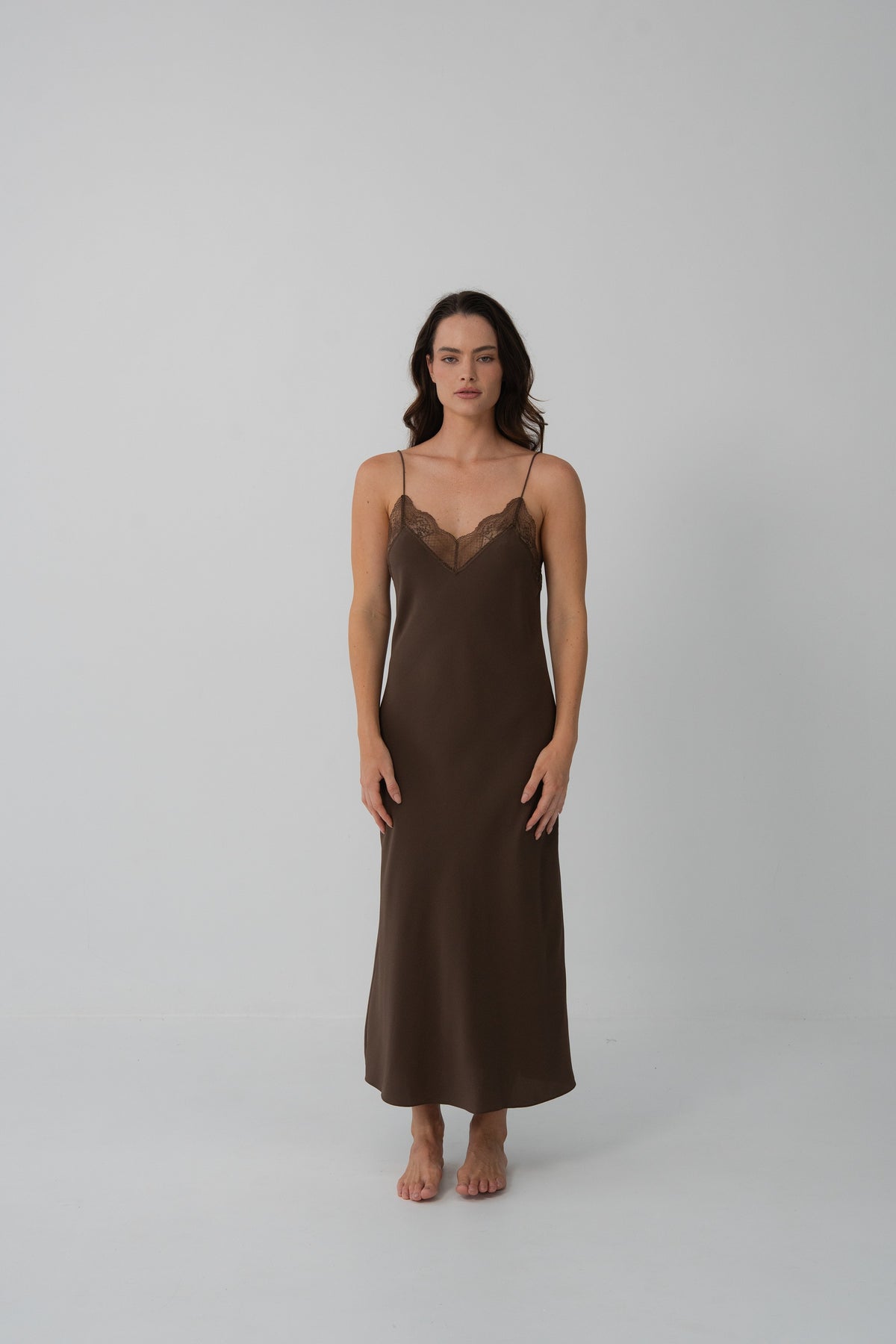 A woman modeling a dark brown slip dress with a lace V-neckline, standing against a plain light gray studio background.
