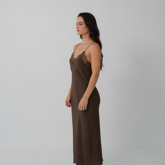 A woman stands in profile wearing a long brown slip dress with thin straps and a lace-trimmed neckline, photographed against a plain light gray studio background.