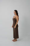 A woman stands in profile wearing a long brown slip dress with thin straps and a lace-trimmed neckline, photographed against a plain light gray studio background.