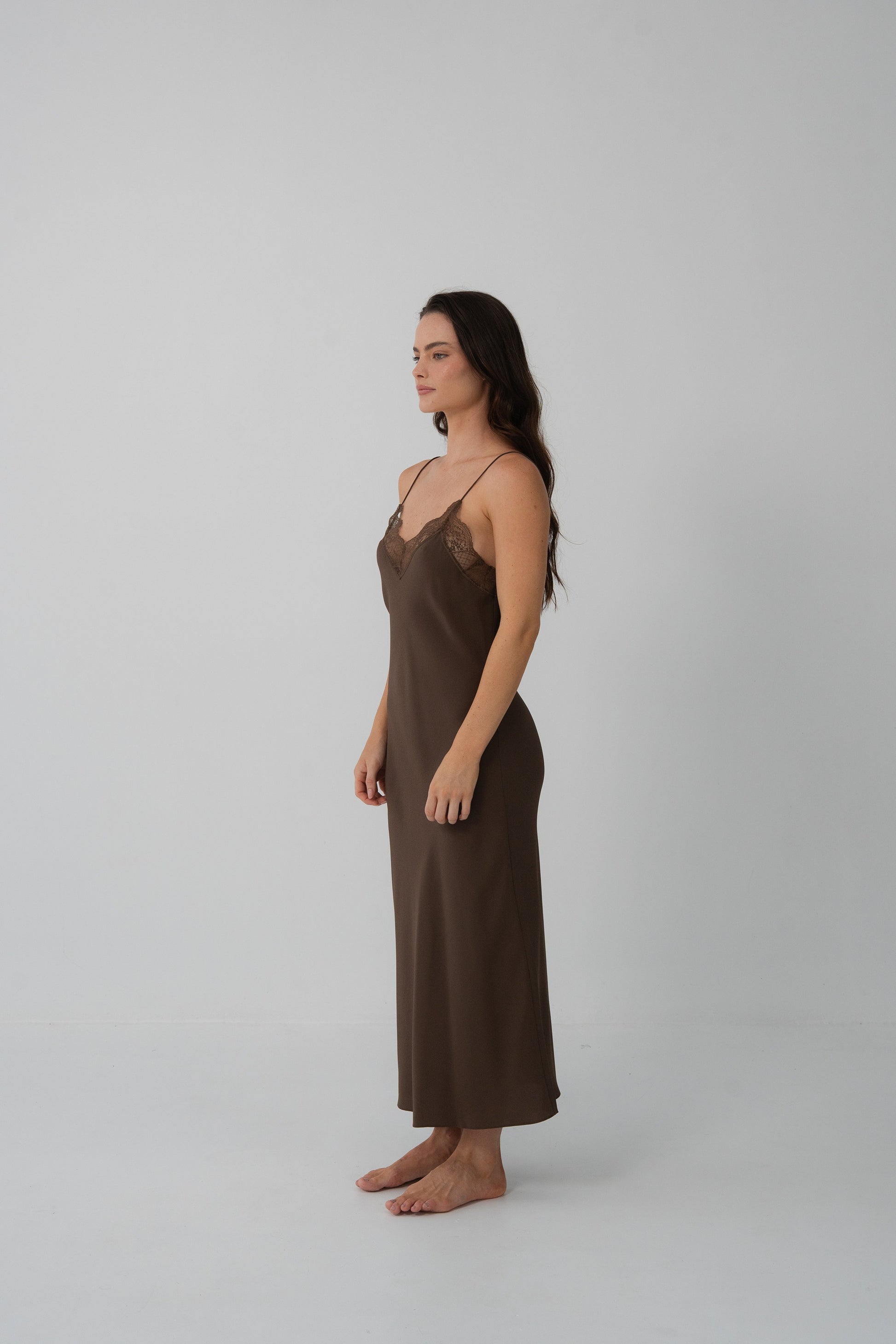 A woman stands in profile wearing a long brown slip dress with thin straps and a lace-trimmed neckline, photographed against a plain light gray studio background.