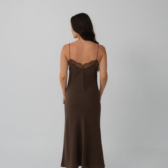 A woman is shown from the back wearing a dark brown slip dress with thin straps and a lace-trimmed neckline, standing against a plain light gray studio background.