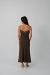 A woman is shown from the back wearing a dark brown slip dress with thin straps and a lace-trimmed neckline, standing against a plain light gray studio background.