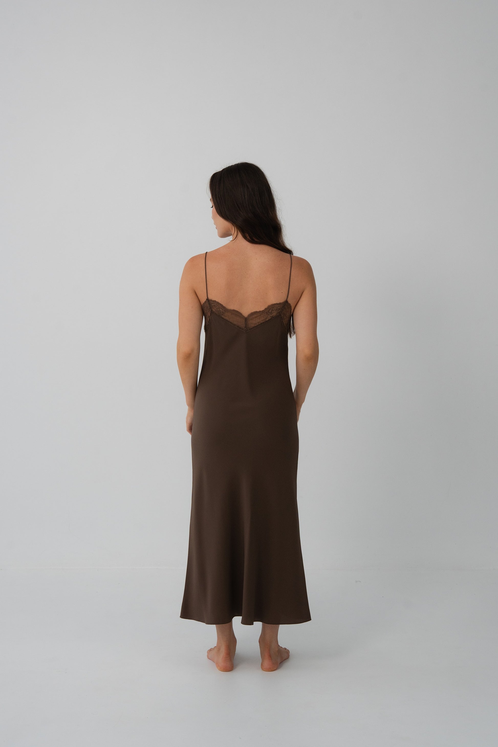A woman is shown from the back wearing a dark brown slip dress with thin straps and a lace-trimmed neckline, standing against a plain light gray studio background.