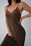 A woman wearing a brown satin slip dress with a lace V-neckline, shown from the shoulders to the hips against a plain light gray studio background.