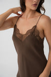 A close-up of a woman wearing a brown satin slip dress with thin straps and a delicate lace V-neckline detail, set against a plain light gray background.