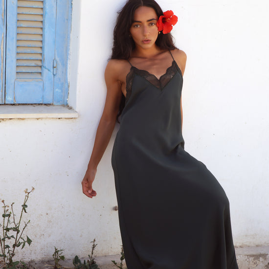 A woman wearing a dark charcoal gray slip dress with black lace trim at the neckline, leaning against a white stucco wall next to a blue shuttered window.