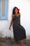A woman wearing a dark charcoal gray slip dress with black lace trim at the neckline, leaning against a white stucco wall next to a blue shuttered window.