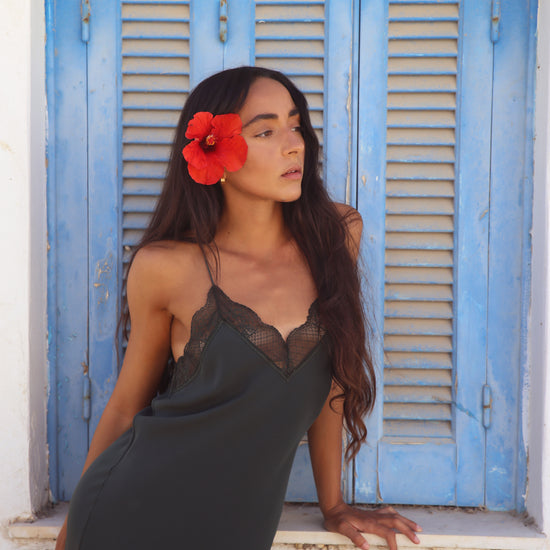 A woman wearing a dark charcoal gray slip dress with black lace trim leans against a white wall in front of a blue shuttered window. She has a bright red flower tucked behind her ear.