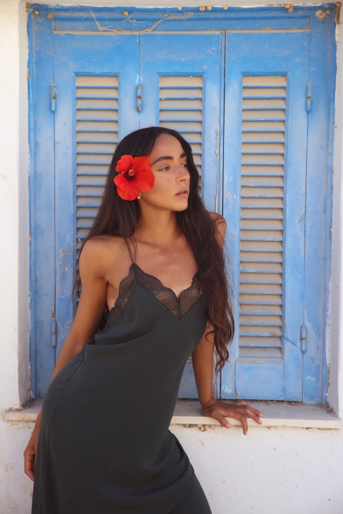 A woman wearing a dark charcoal gray slip dress with black lace trim leans against a white wall in front of a blue shuttered window. She has a bright red flower tucked behind her ear.