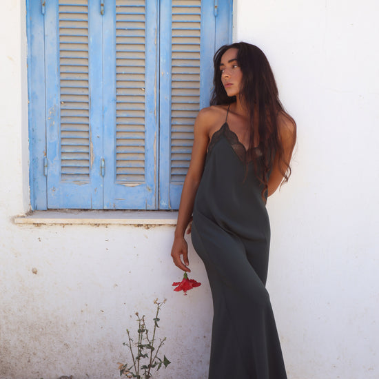 A woman wearing a dark gray slip dress with lace trim stands barefoot against a white wall next to a blue shuttered window, holding a small red flower.