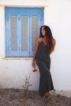 A woman wearing a dark gray slip dress with lace trim stands barefoot against a white wall next to a blue shuttered window, holding a small red flower.