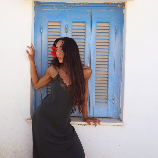 A woman with long dark hair wearing a black slip dress leans against a white wall next to a blue shuttered window with a terracotta tile trim.