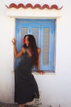 A woman with long dark hair wearing a black slip dress leans against a white wall next to a blue shuttered window with a terracotta tile trim.