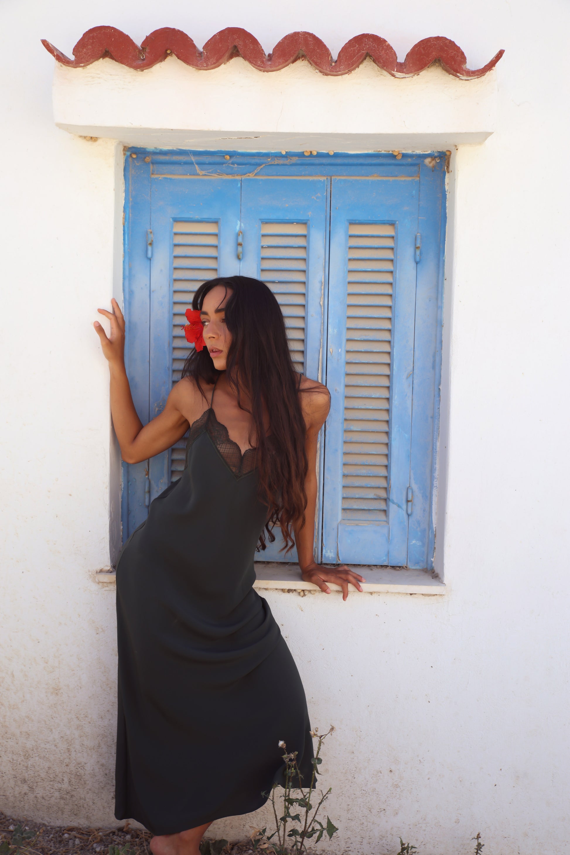A woman with long dark hair wearing a black slip dress leans against a white wall next to a blue shuttered window with a terracotta tile trim.
