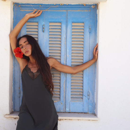 A woman with long dark hair poses in front of a white wall and a blue shuttered window, wearing a dark gray sleeveless slip dress and holding a red flower.