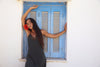 A woman with long dark hair poses in front of a white wall and a blue shuttered window, wearing a dark gray sleeveless slip dress and holding a red flower.