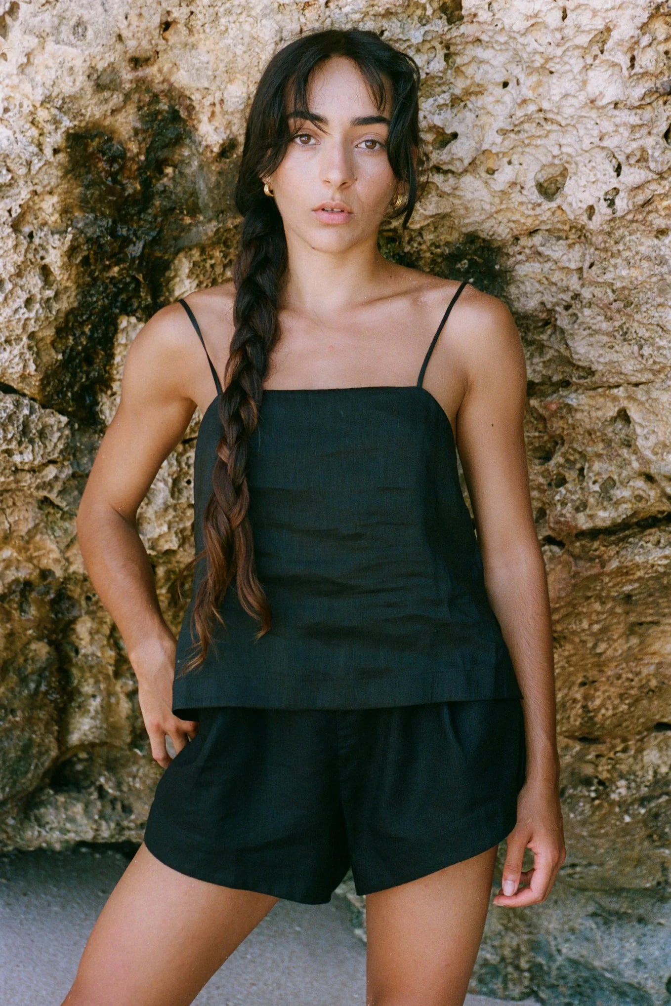 Woman wearing a black tank top and shorts against a stone wall.