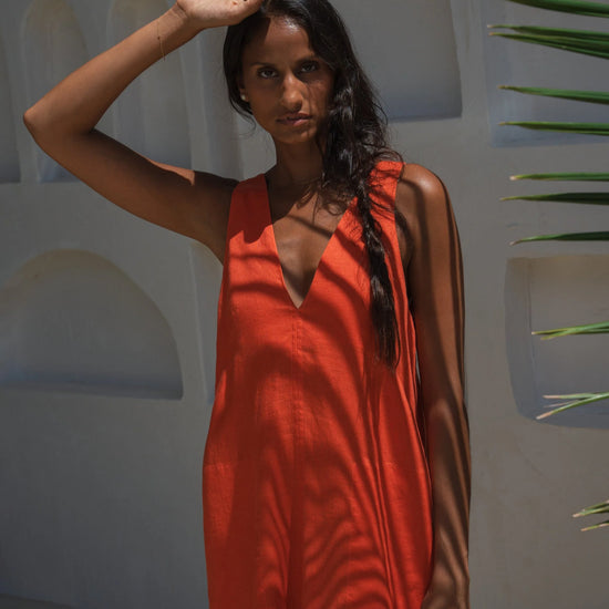 Woman in an orange linen dress standing against a white wall with greenery.
