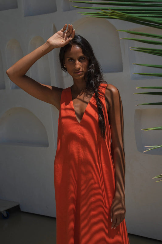 Woman in an orange linen dress standing against a white wall with greenery.