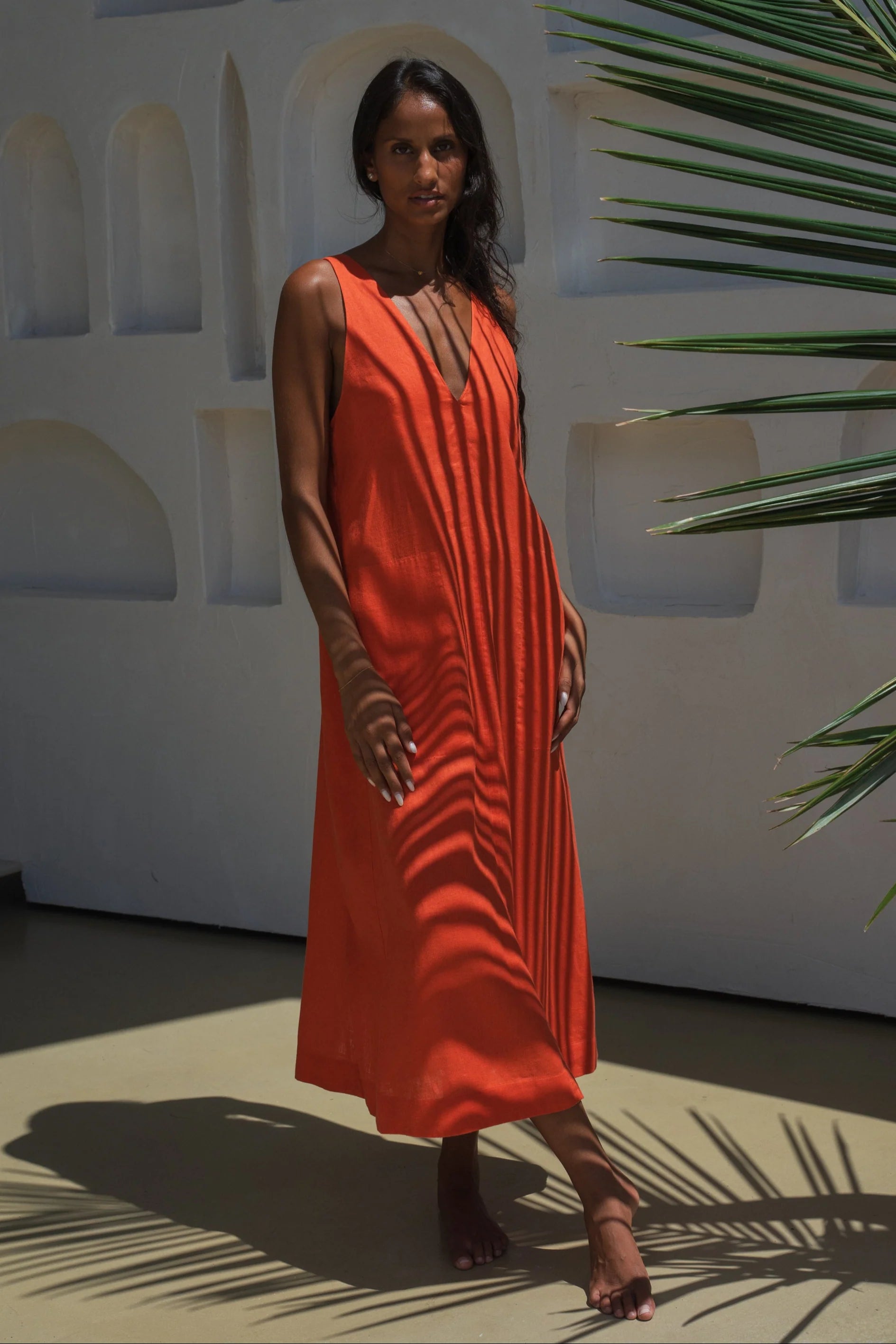 Woman in an orange linen dress standing against a white wall with architectural elements and greenery.