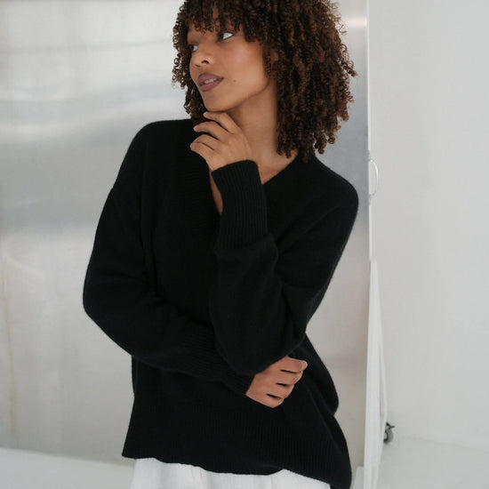 Woman wearing a black sweater and white pants against a neutral background