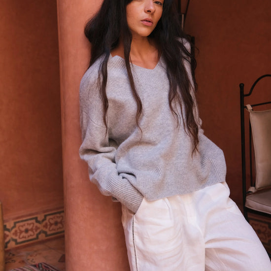 Woman wearing a gray sweater and white pants in a warm-toned interior setting