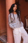 Woman wearing a gray sweater and white pants in a warm-toned interior setting