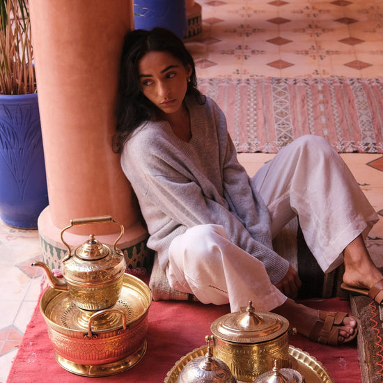 A woman in a light gray robe and white pants sits on a red floor mat in a Moroccan-style courtyard, surrounded by brass and silver tea sets and decorative columns.